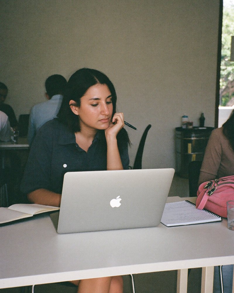 Desarrolladora web mexicana de unos veinte años pensativa en una mesa de coworking, con laptop y libreta
