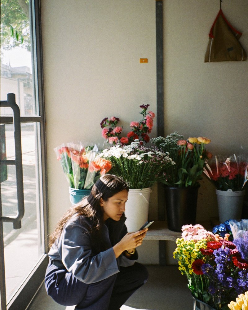 Joven dueña de una floristería en México se pone en cuclillas junto a cubetas de flores mientras revisa su celular