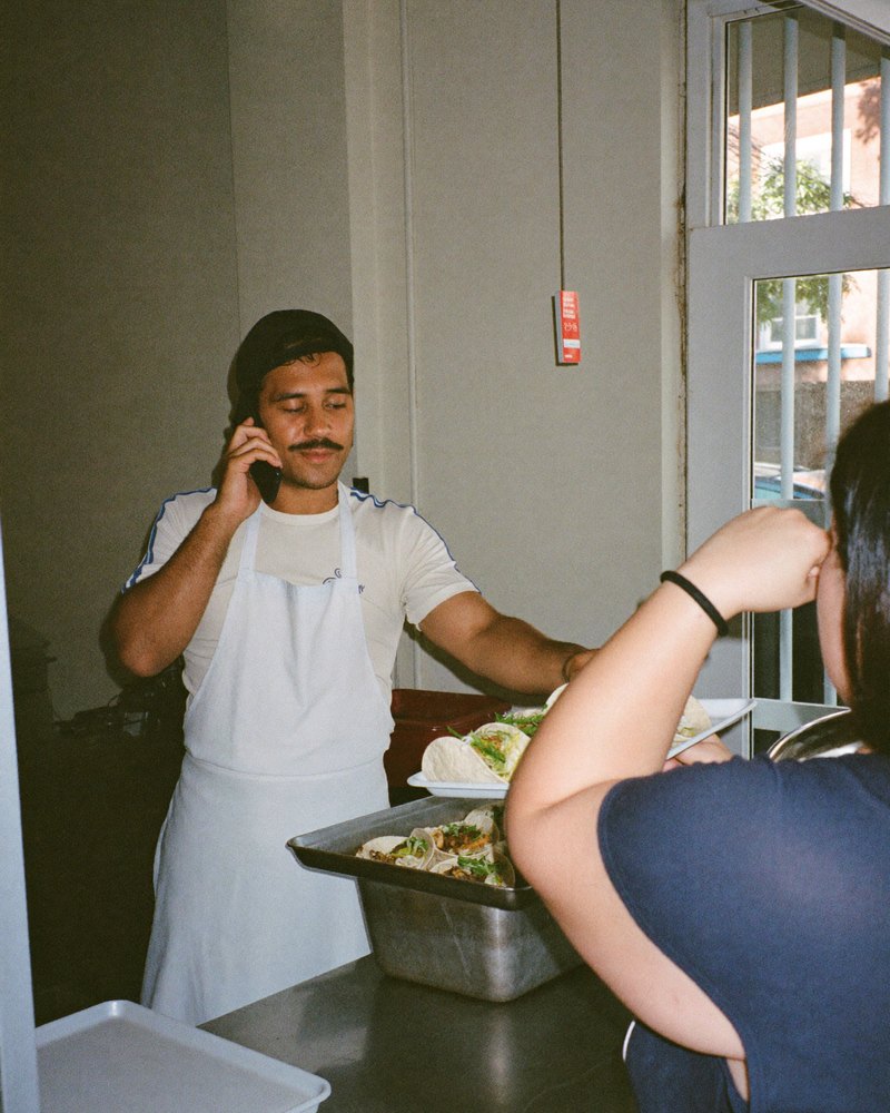 Taquero con delantal en su puesto de tacos mostrando su celular a un cliente